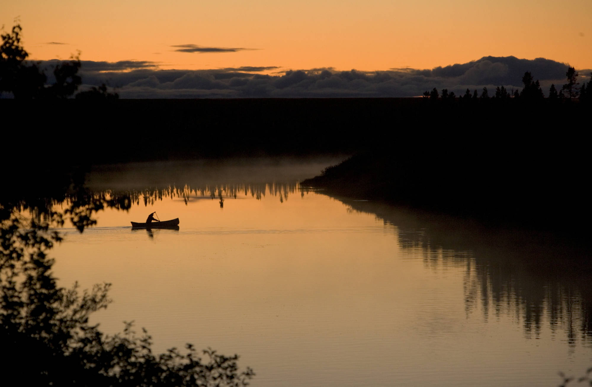 在日出或日落时，划桨者在平静的湖面上划独木舟的剪影