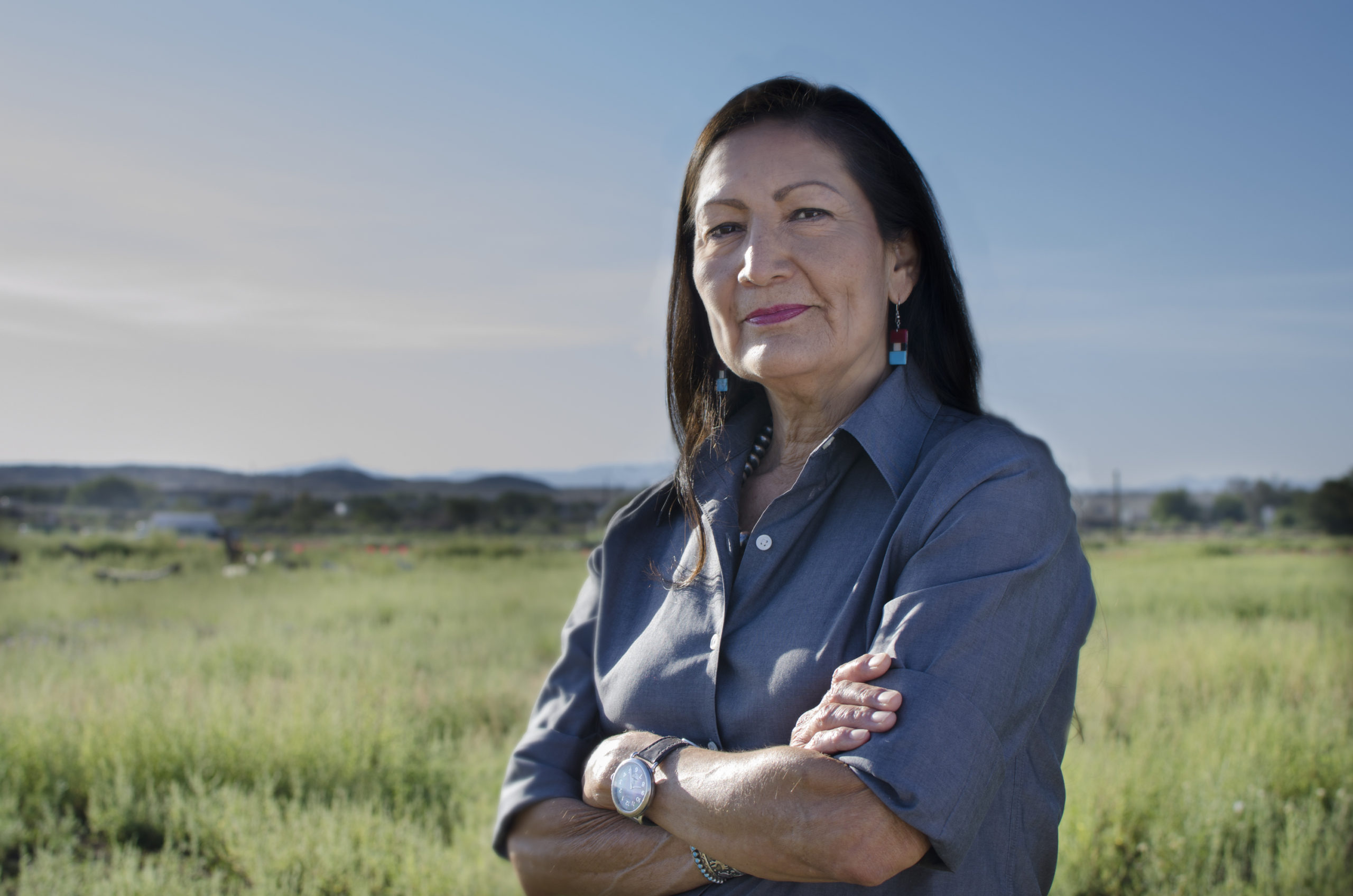 一位深色头发、戴着绿松石耳环的新墨西哥妇女交叉双臂，在开阔的田野里看着镜头。