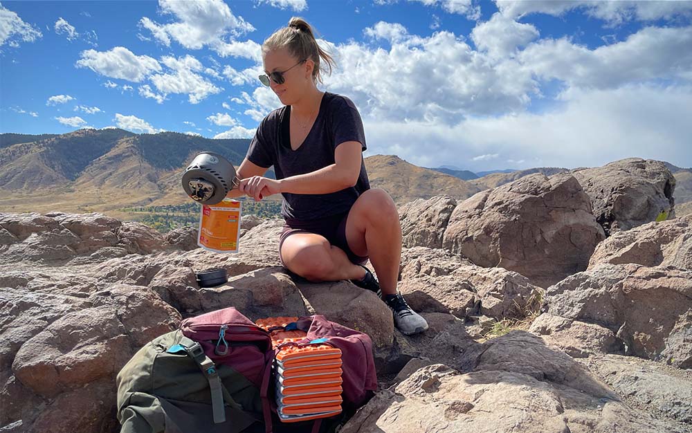 一名妇女坐在一块俯瞰群山的岩石上，把水倒进一袋脱水的橙色食物里