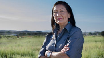 A New Mexican woman with dark hair and turquoise earrings crosses her arms and looks at the camera in an open field.